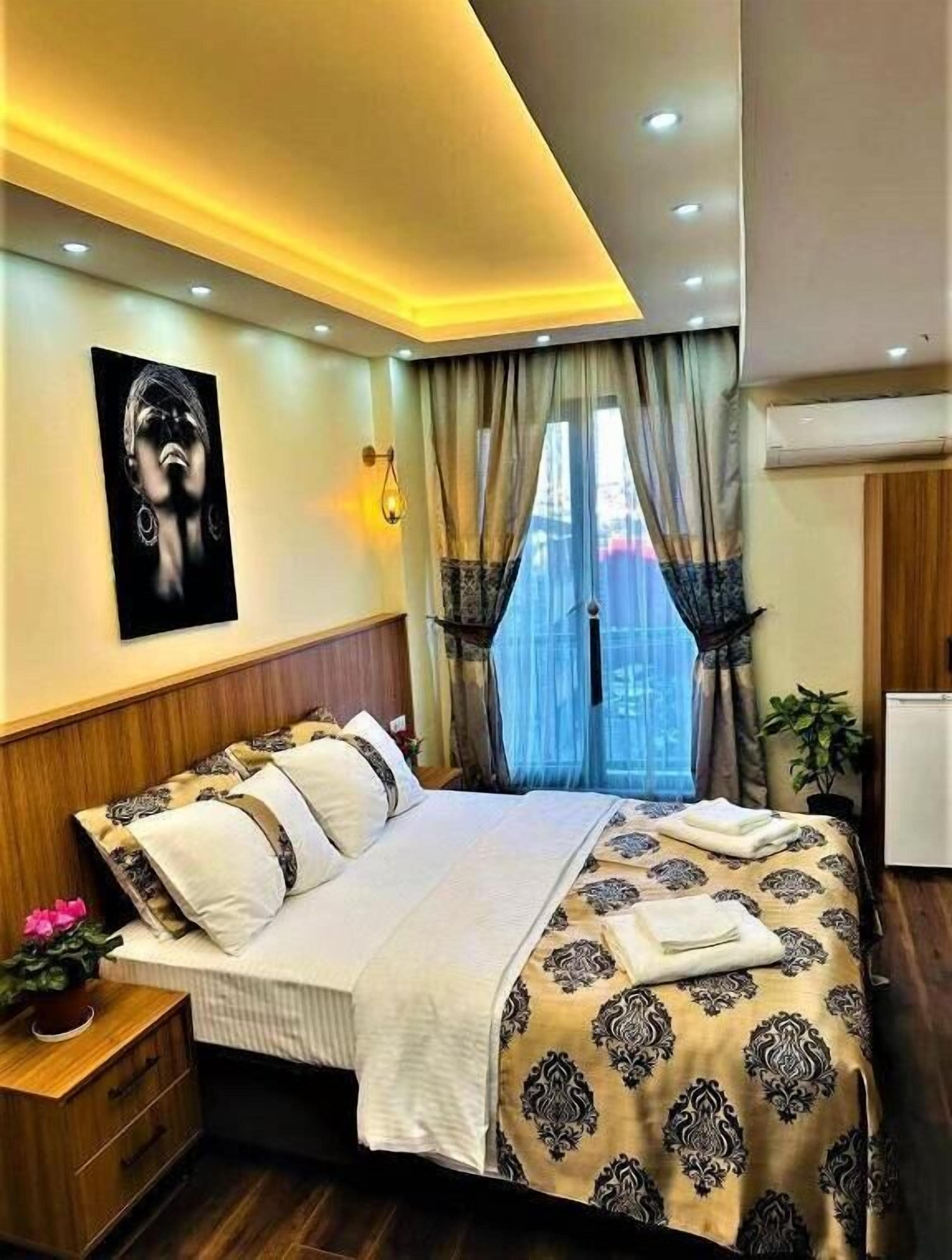 Une chambre à coucher comprenant un lit, une table de chevet, une œuvre d’art accrochée au mur, une fenêtre avec des rideaux et un appareil de climatisation.