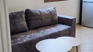 Ett vardagsrum med en grå soffa, ett litet vitt bord, ett fönster med utsikt över grönområden och en köksdel med ett kylskåp.
