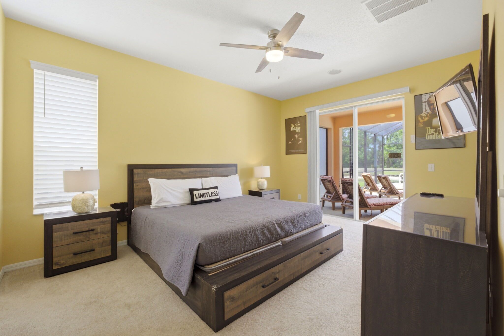 A bedroom with a bed, nightstands, a ceiling fan, a window with blinds, and a view of outdoor furniture through a glass door.