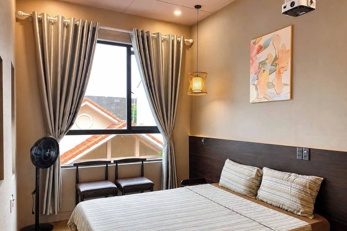 A bedroom with a bed, a desk, a chair, a standing fan, and a window with curtains.