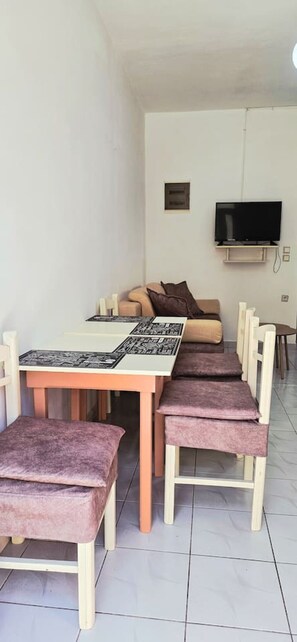 Ein Essbereich mit einem langen Tisch, gepolsterten Stühlen, einem an der Wand befestigten Fernseher und einer Couch im Hintergrund.
