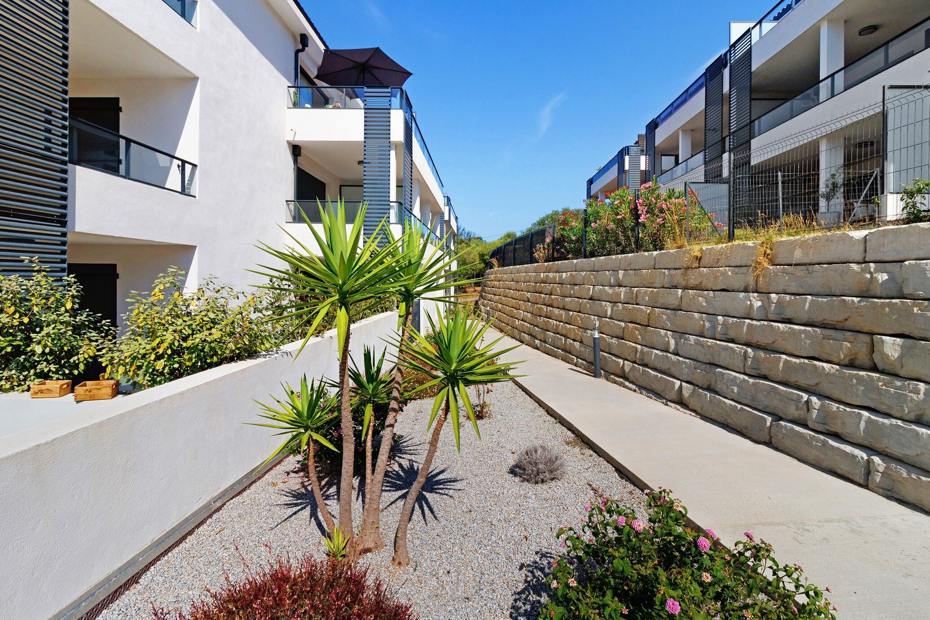 A modern building with balconies, a stone retaining wall, and a variety of plants.