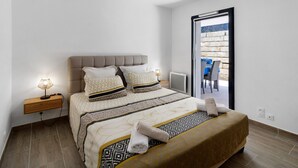 Una cama bien tendida con una colcha estampada, una lámpara de noche y vista a un área de estar exterior a través de un ventanal.