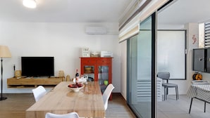 Un salon moderne avec une table à manger en bois, des chaises blanches, un téléviseur à écran plat, un meuble rouge et une porte-fenêtre coulissante donnant sur un balcon.