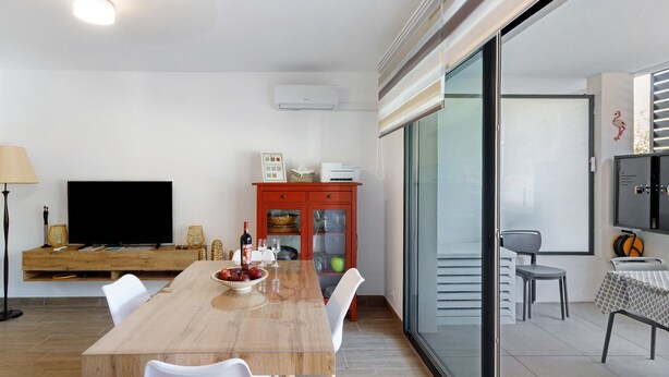 Una sala de estar moderna con una mesa de comedor de madera, sillas blancas, un televisor de pantalla plana, un armario rojo y una puerta de vidrio corredera que da a un balcón.