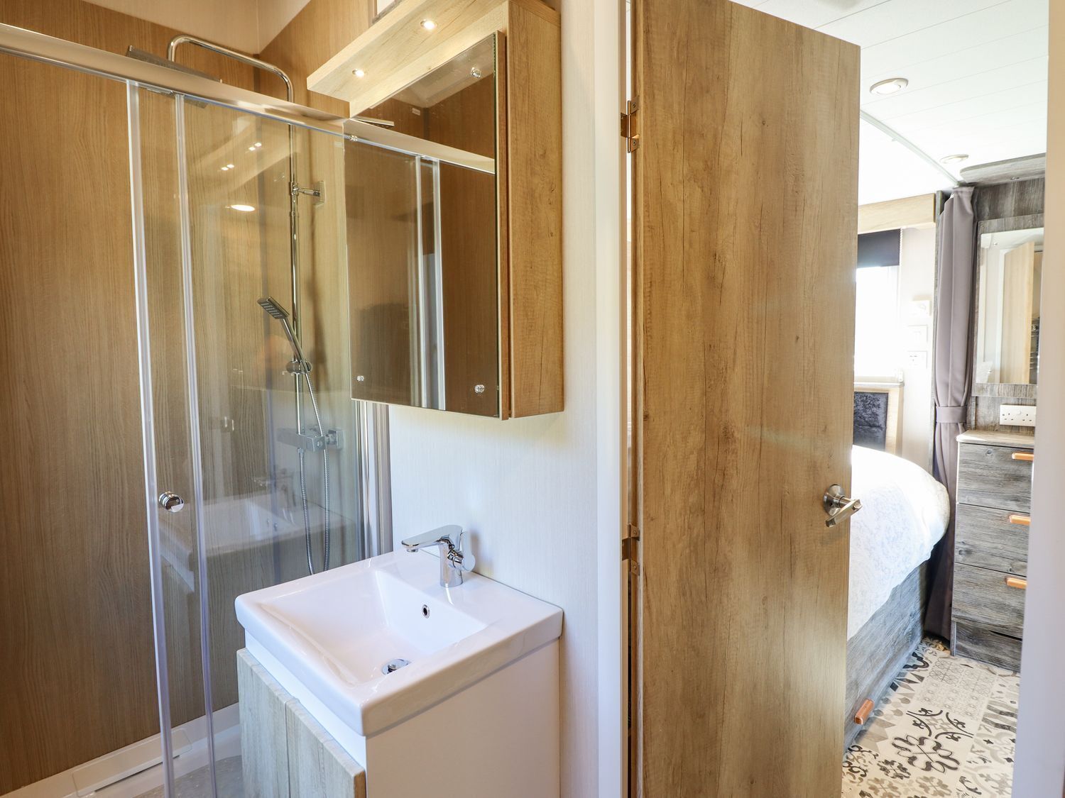A modern bathroom with a glass shower enclosure, a white sink, and a wooden door leading to a bedroom.
