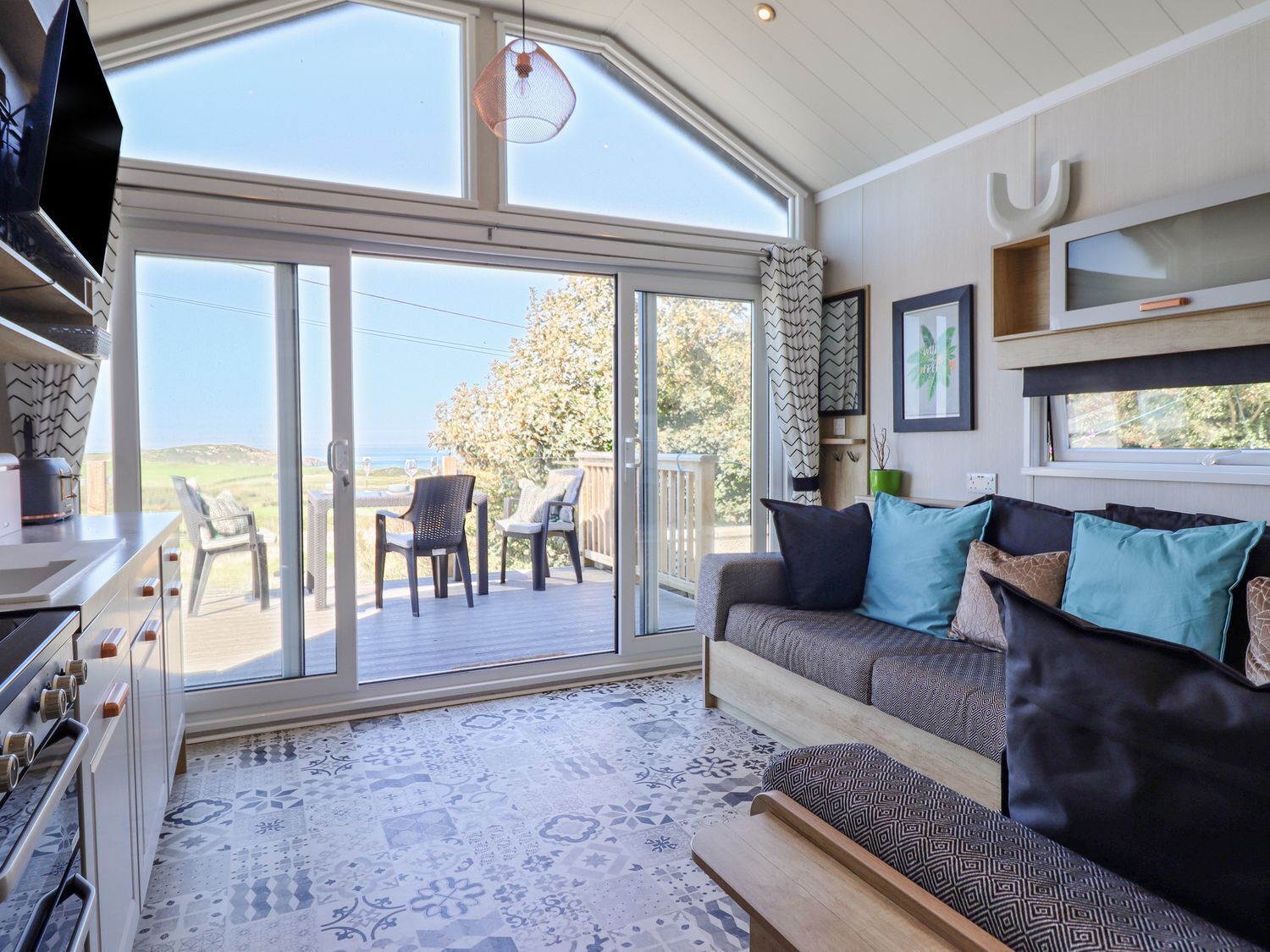 A modern living room with a large sliding glass door leading to an outdoor deck, a sofa with patterned cushions, a television, and a kitchen area with a stove.