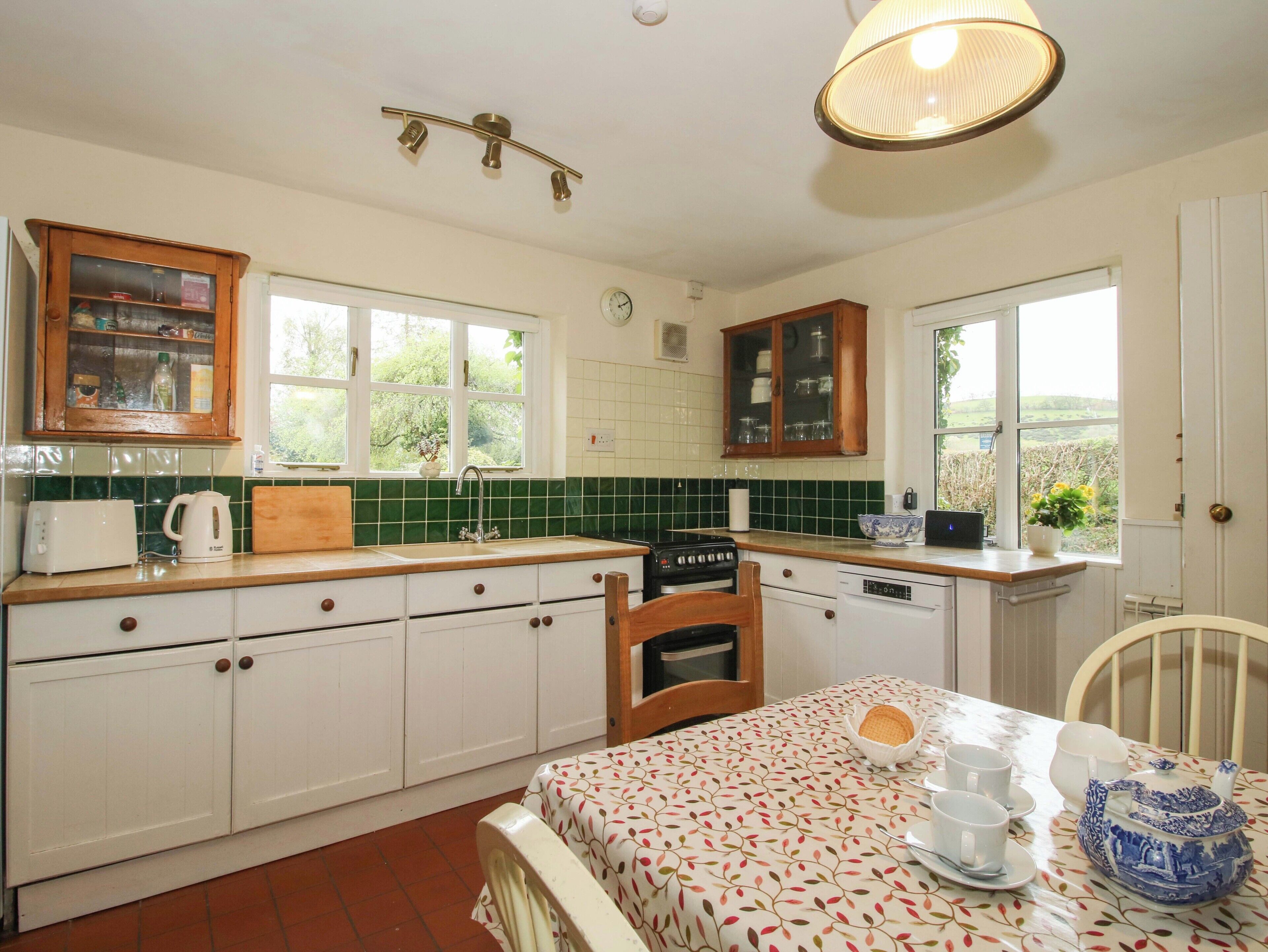 A kitchen with a sink, stove, and cabinets, a dining table with tea set, and a window with a view of greenery.