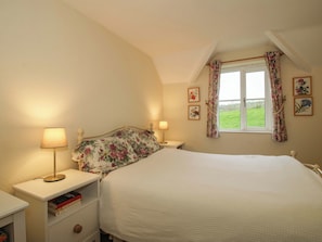 A bedroom with a bed, two bedside tables, a window with curtains, and a view of a green field. - Ivy Cottage (Church Stretton)