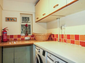 A laundry area with a washing machine, a fire extinguisher, a wicker basket, and a window with a decorative item. - Ivy Cottage (Church Stretton)