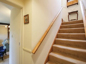 Una escalera con alfombra y barandillas de madera, un cuadro enmarcado en la pared, y una vista parcial de una sala con un sillón azul.