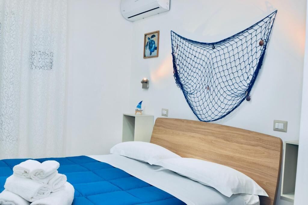 A hotel room with a bed, a blue quilted bedspread, white pillows, a wooden headboard, a wall-mounted hammock, a small table with a lamp, and a framed picture on the wall.