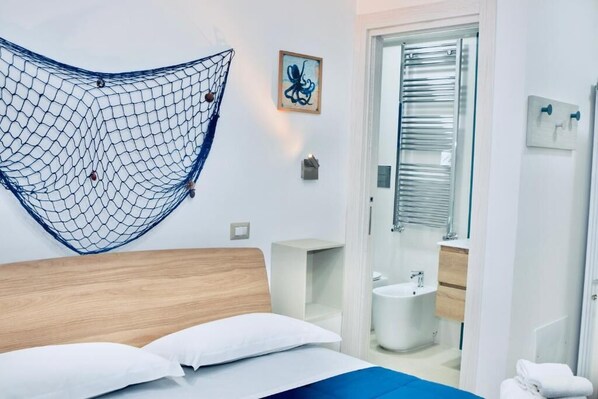 Habitación de hotel moderna con cama, hamaca, baño con lavamanos y espejo, y una puerta que da a otra habitación.
