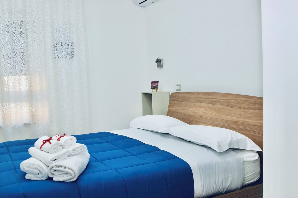Una cama bien hecha con colcha azul, almohadas blancas y toallas dobladas sobre un cabecero blanco.