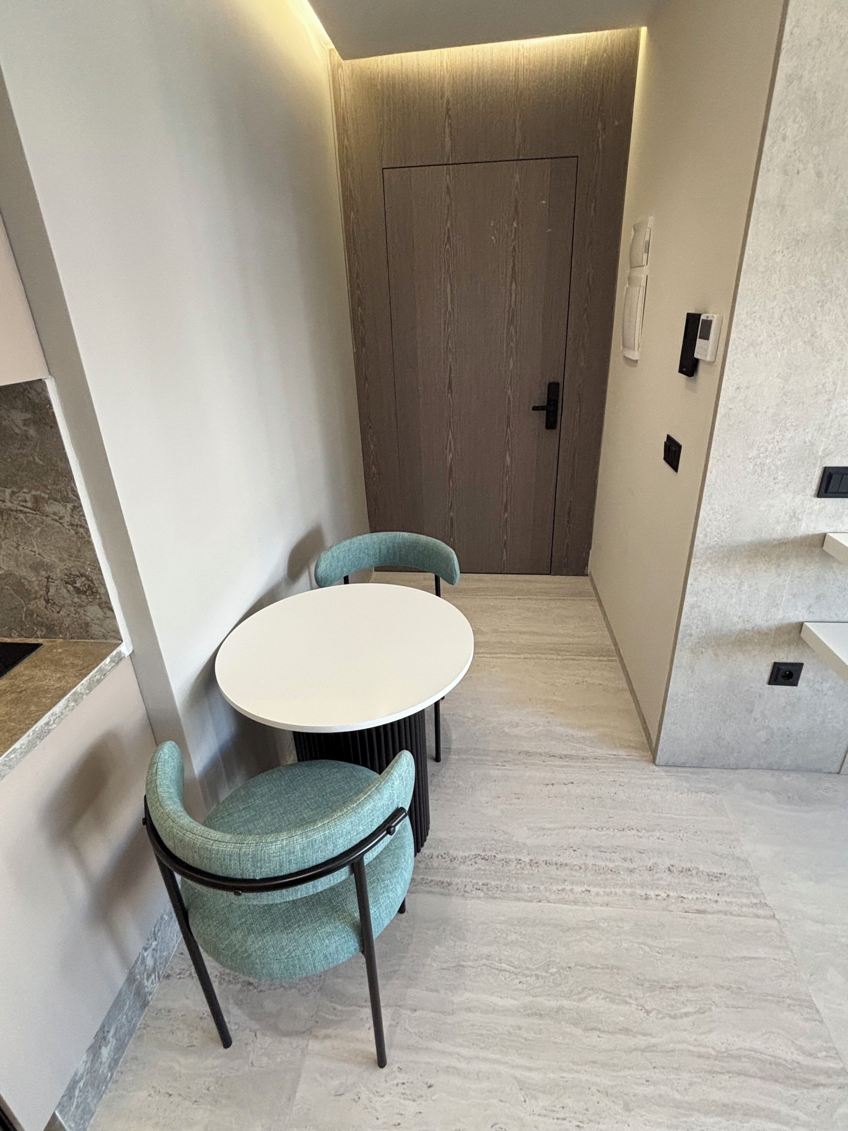 Une pièce d’angle avec une table ronde et deux chaises, une porte en bois et un mur blanc avec un interrupteur.
