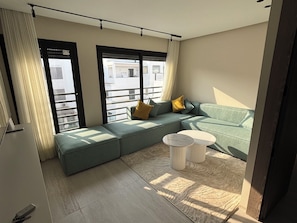 Ein modernes Wohnzimmer mit einer Eckcouch, einem runden Couchtisch und großen Fenstern mit Jalousien.
