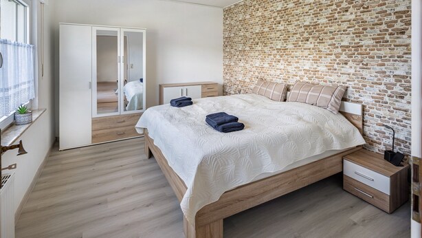 A bedroom with a brick wall, wooden bed frame, and a bedside table with a lamp.