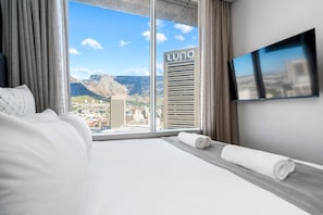A hotel room with a large window, a bed, a flat-screen TV, and a view of a cityscape and mountains. - One Thibault 1927 (Cape Town)