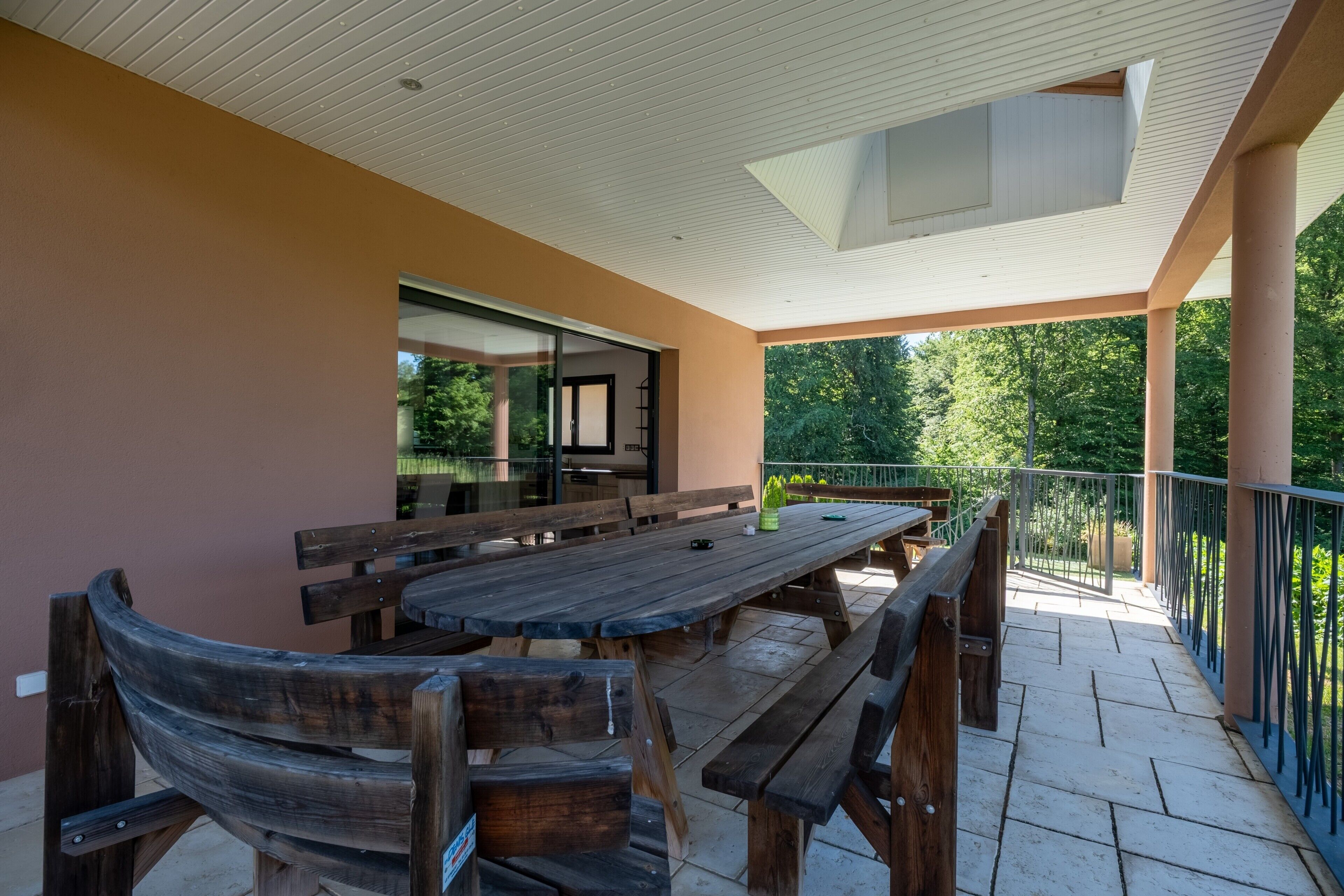 Eine überdachte Terrasse mit einem Holztisch und Bänken, eine Glastür, die zu einem Innenbereich führt, und ein Balkon mit Geländer und Blick auf Grünflächen.