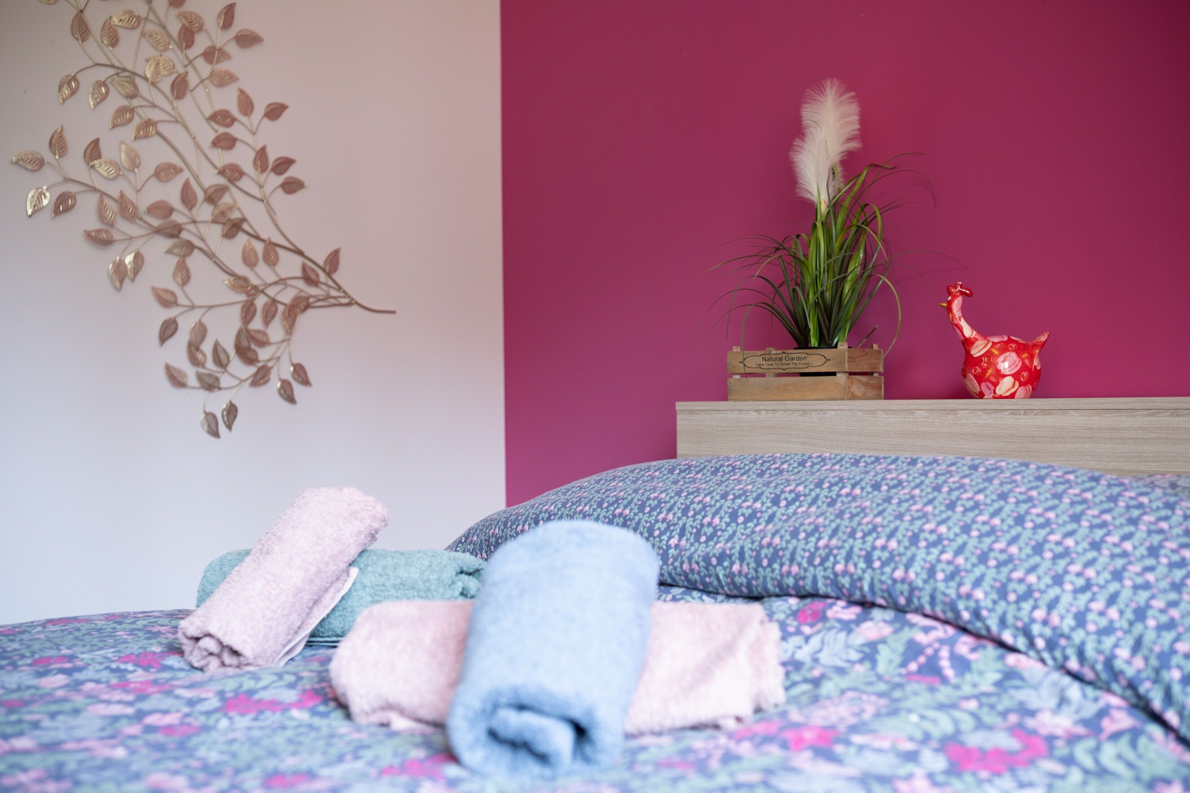 Une chambre avec une couvre-lit à motifs floraux, un élément décoratif mural, une plante et un petit vase rouge.