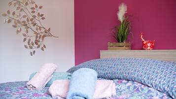 Une chambre avec une couvre-lit à motifs floraux, un élément décoratif mural, une plante et un petit vase rouge.