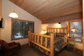 Une chambre à coucher avec un lit en bois, une grande fenêtre avec des stores, une chaise et une table de chevet avec une lampe.