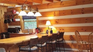 A cabin interior with a wooden interior, a kitchen area with hanging pots, a dining area with chairs, and a bar counter. - Peaceful 2-bedroom cabin with AC in charming Logan Sleeps 8 (Logan)