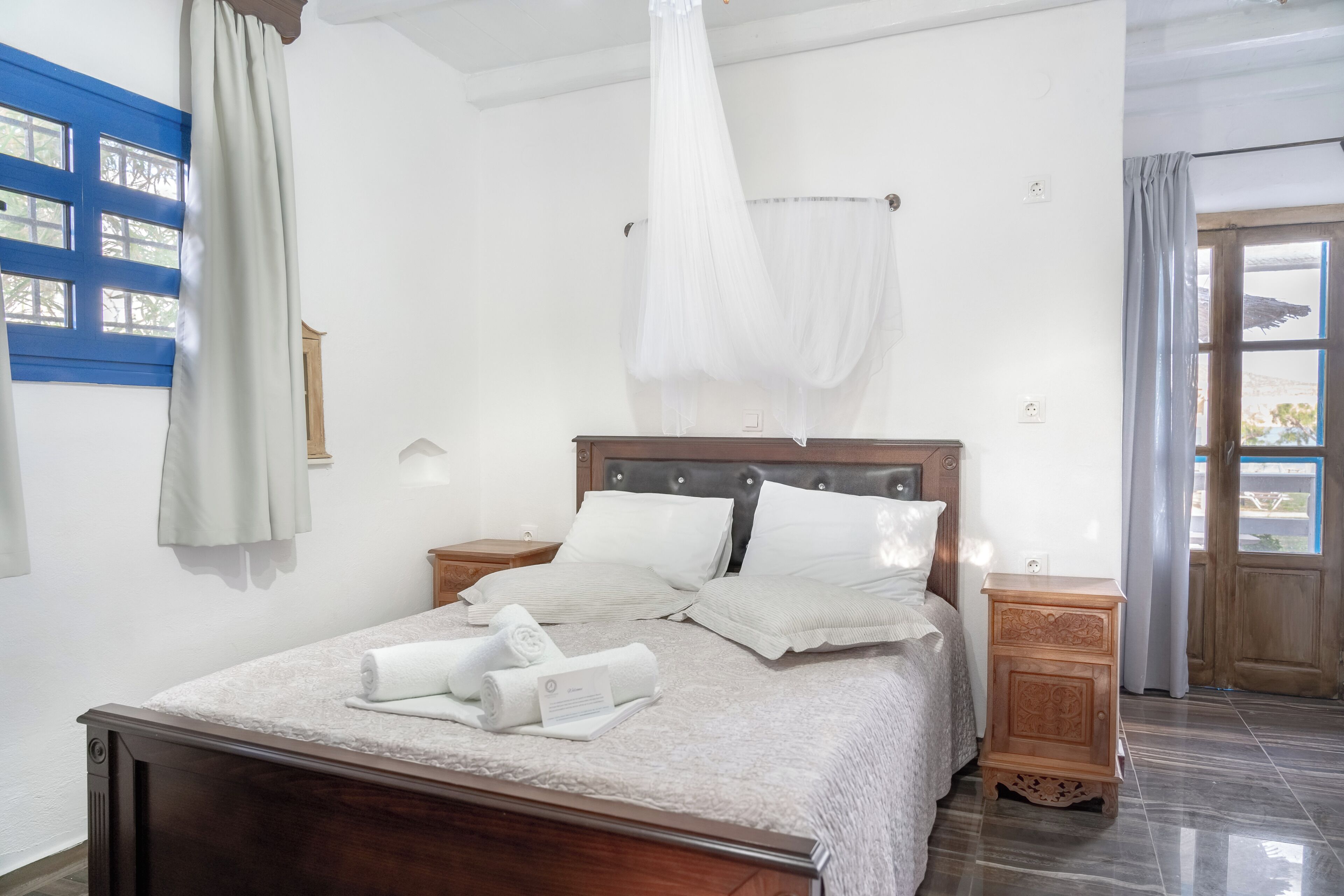 A neatly made bed with pillows and a rolled towel, a bedside table, a nightstand, and a window with curtains.