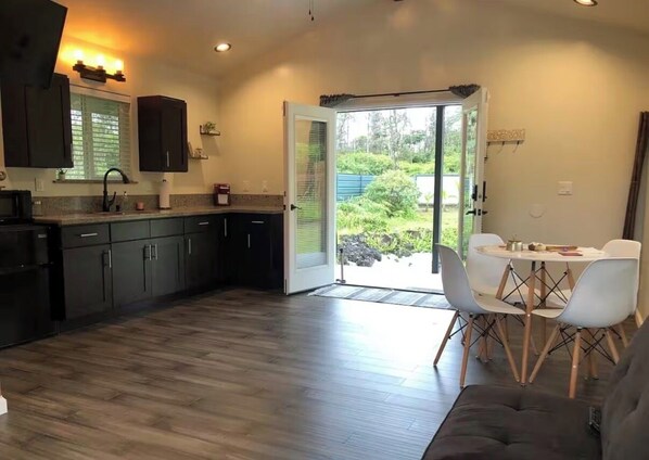 A modern kitchen with dark cabinets, a countertop, and a dining area with white chairs and a table. - Great for nature lovers quiet and calm. Auto gat, security cameras. (Keaau)