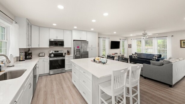 A modern kitchen with white cabinetry, a central island, and a dining area with a gray sofa and white chairs. - Creature Comforts (Rehoboth Beach)