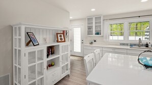 A modern kitchen with white cabinetry, a dining area, and a large window with a view of greenery. - Creature Comforts (Rehoboth Beach)