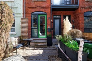 A brick building with a green door, a small outdoor seating area with a bench and table, and a planter with plants.