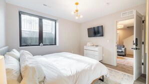 A bedroom with a bed, a TV, a window, a dresser, and a door leading to another room. - Private 3BR Residence | Modern Condo near U of M Stadium | Sleeps 6 (Ann Arbor)