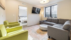 A modern living room with a grey sofa, green armchairs, and a small ottoman. A flat-screen TV is mounted on the wall, and a window provides natural light. - Private 3BR Residence | Modern Condo near U of M Stadium | Sleeps 6 (Ann Arbor)