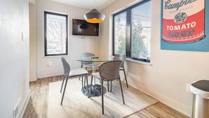 A dining area with a glass table, metal chairs, a hanging pendant light, and a large poster of Campbell's Tomato Soup. - Private 3BR Residence | Modern Condo near U of M Stadium | Sleeps 6 (Ann Arbor)