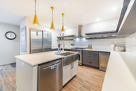 A modern kitchen with stainless steel appliances, a dishwasher, and a sink. There are yellow pendant lights, a clock on the wall, and a tiled backsplash.
