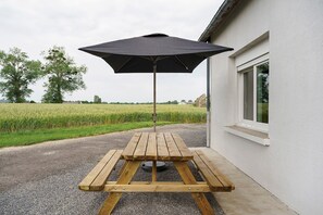 A wooden picnic table with an umbrella, a gravel path, a white building, and a field in the background.