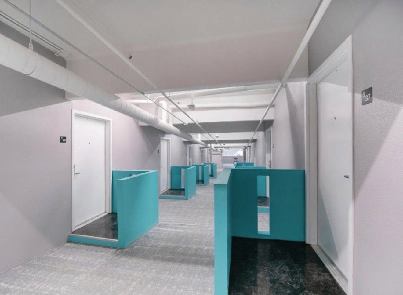 Un pasillo con cubículos azul turquesa, puertas blancas y suelo de baldosas.