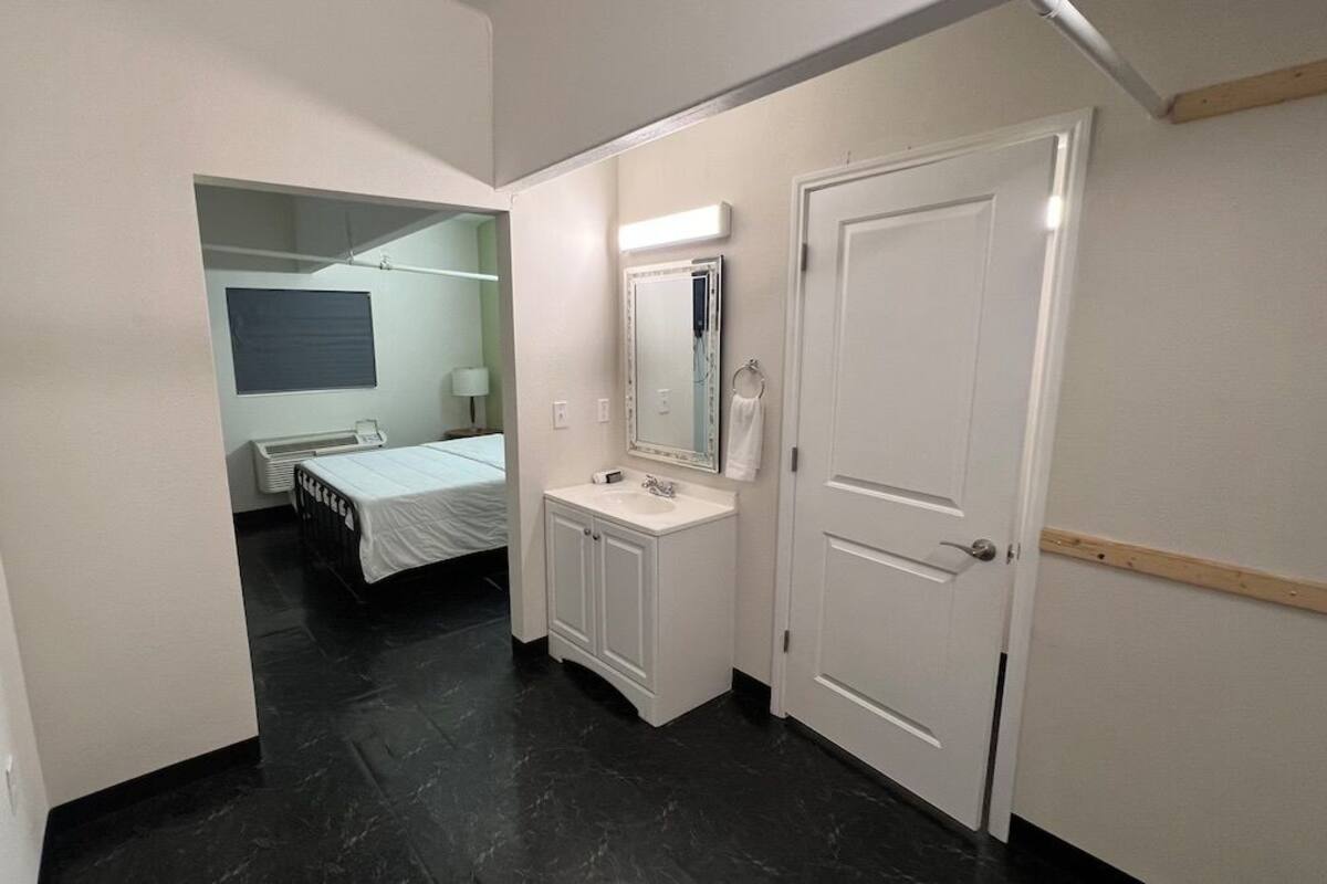 Un pasillo que conduce a un dormitorio con cama, un baño con lavamanos y espejo, y una puerta.
