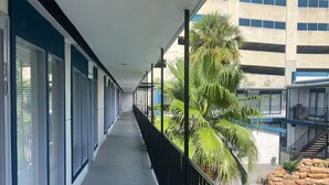 A long corridor with a pool area on one side, featuring a concrete floor, metal railings, and a view of a multi-story building with windows.