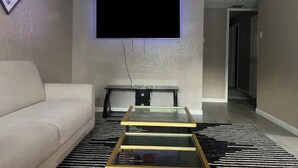 A living room with a sofa, a coffee table, a flat-screen TV mounted on the wall, a bench, and a patterned rug.
