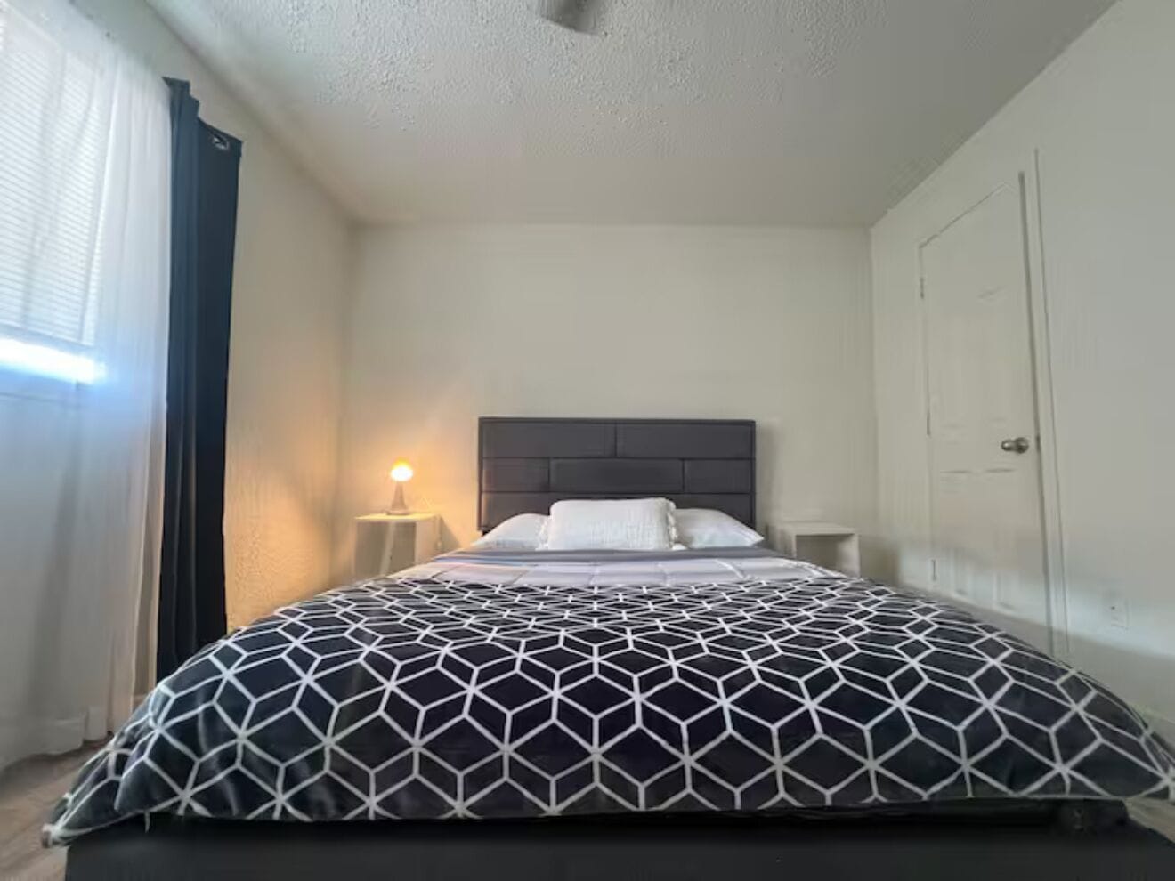 Un dormitorio con cama, mesita de noche, lámpara, puerta y ventana con cortinas.