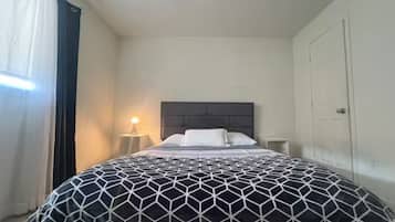 Un dormitorio con cama, mesita de noche, lámpara, puerta y ventana con cortinas.