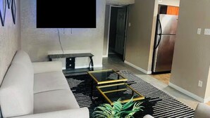 A living room with a sofa, a flat-screen TV mounted on the wall, a coffee table, and a kitchen area in the background.