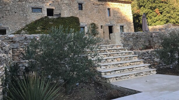 Edificio in pietra con una scala che conduce a una zona pianeggiante, circondato da vegetazione.