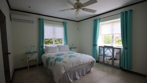 Ein Schlafzimmer mit einem Bett, einem Deckenventilator, einem Fenster mit Vorhängen, einem Nachttisch und einem kleinen Tisch.