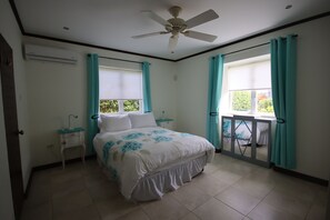 Un dormitorio con cama, ventilador de techo, ventana con cortinas, mesita de noche y una pequeña mesa.