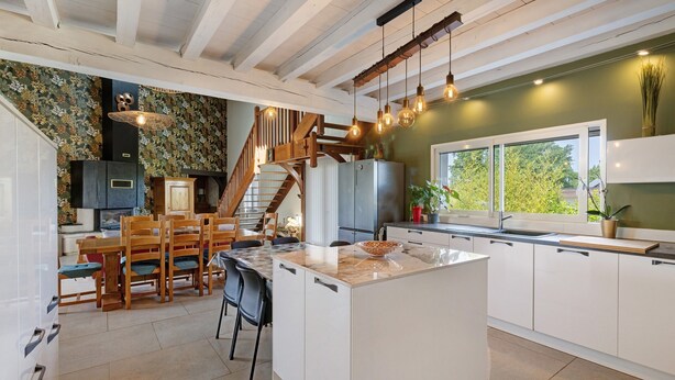 A spacious kitchen with a central island, wooden chairs, a staircase, and a large window.
