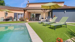 A swimming pool with lounge chairs and umbrellas, a patio area, and a building with a tiled roof.