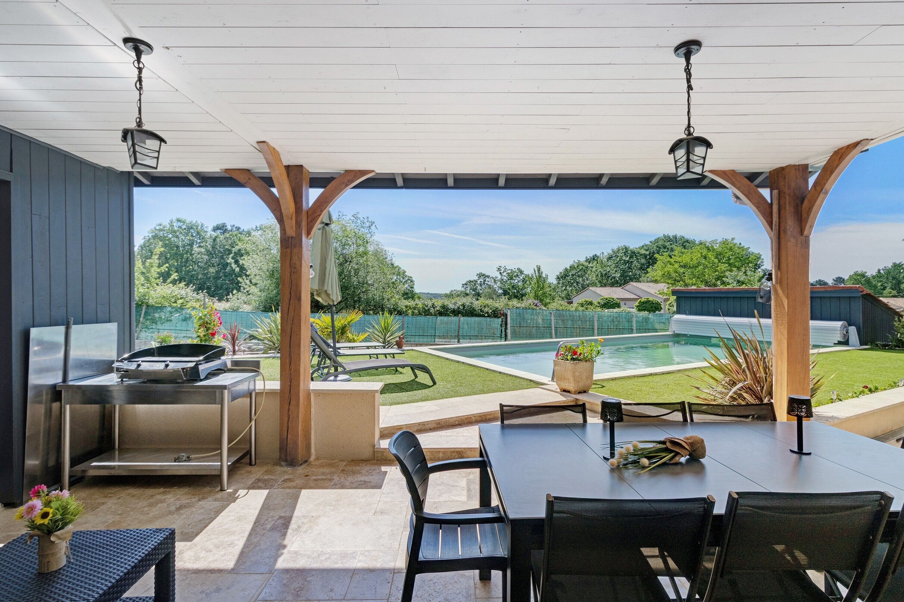 Une terrasse extérieure équipée d’un barbecue, d’une table à manger, de chaises et offrant une vue sur une piscine et un jardin.